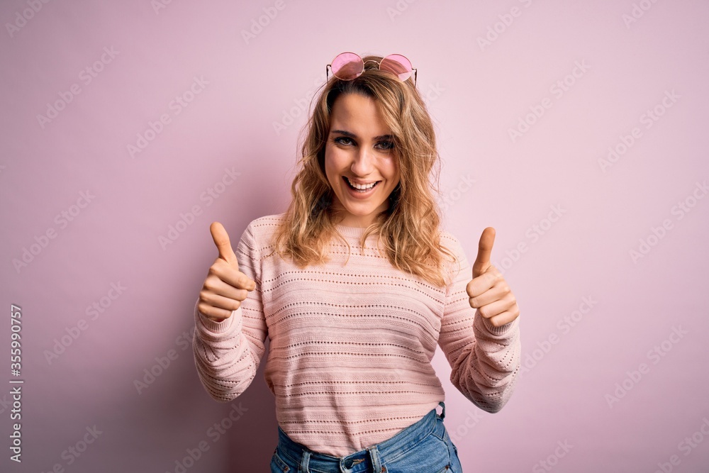 Young beautiful blonde woman wearing casual sweater and sunglasses over pink background success sign doing positive gesture with hand, thumbs up smiling and happy. Cheerful expression and winner