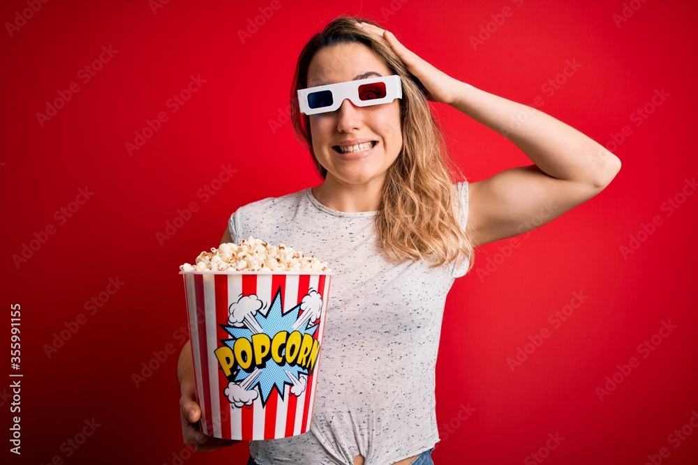 Obraz premium Young beautiful blonde woman watching movie using 3d glasses eating popcorn snack stressed with hand on head, shocked with shame and surprise face, angry and frustrated. Fear and upset for mistake.