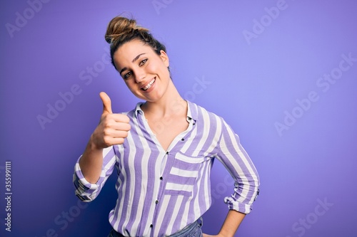 Young beautiful blonde woman wearing casual striped shirt standing over purple background doing happy thumbs up gesture with hand. Approving expression looking at the camera showing success.