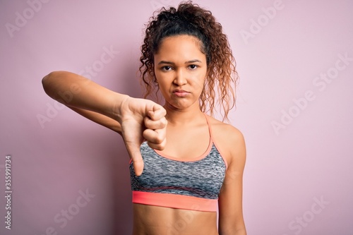 Beautiful sportswoman with curly hair doing sport wearing sportswear over pink background looking unhappy and angry showing rejection and negative with thumbs down gesture. Bad expression. © Krakenimages.com