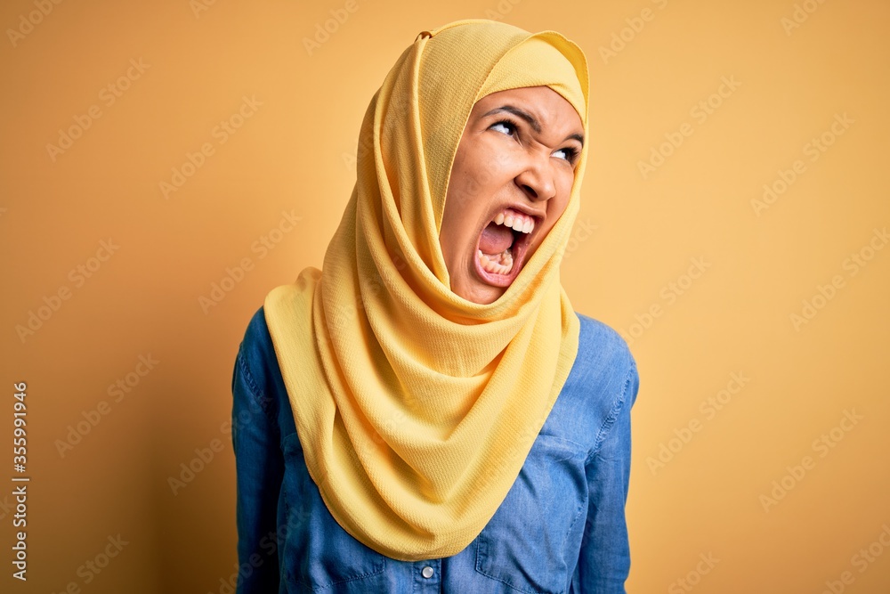 Young beautiful woman with curly hair wearing arab traditional hijab over yellow background angry and mad screaming frustrated and furious, shouting with anger. Rage and aggressive concept. Young beautiful woman with curly hair wearing arab traditional hijab over yellow background angry and mad screaming frustrated and furious, shouting with anger. Rage and aggressive concept.