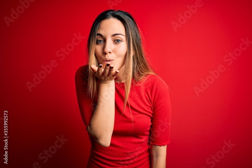 Young beautiful blonde woman with blue eyes wearing casual t-shirt over red background looking at the camera blowing a kiss with hand on air being lovely and sexy. Love expression.