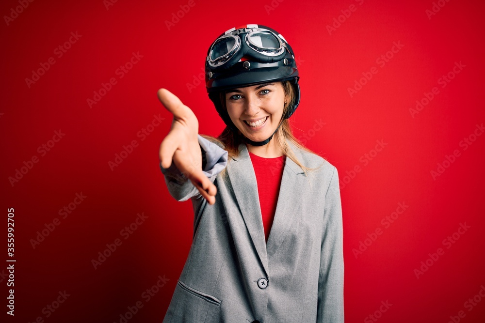 Young beautiful blonde motorcyclist woman wearing motorcycle helmet over red background smiling friendly offering handshake as greeting and welcoming. Successful business. Young beautiful blonde motorcyclist woman wearing motorcycle helmet over red background smiling friendly offering handshake as greeting and welcoming. Successful business.