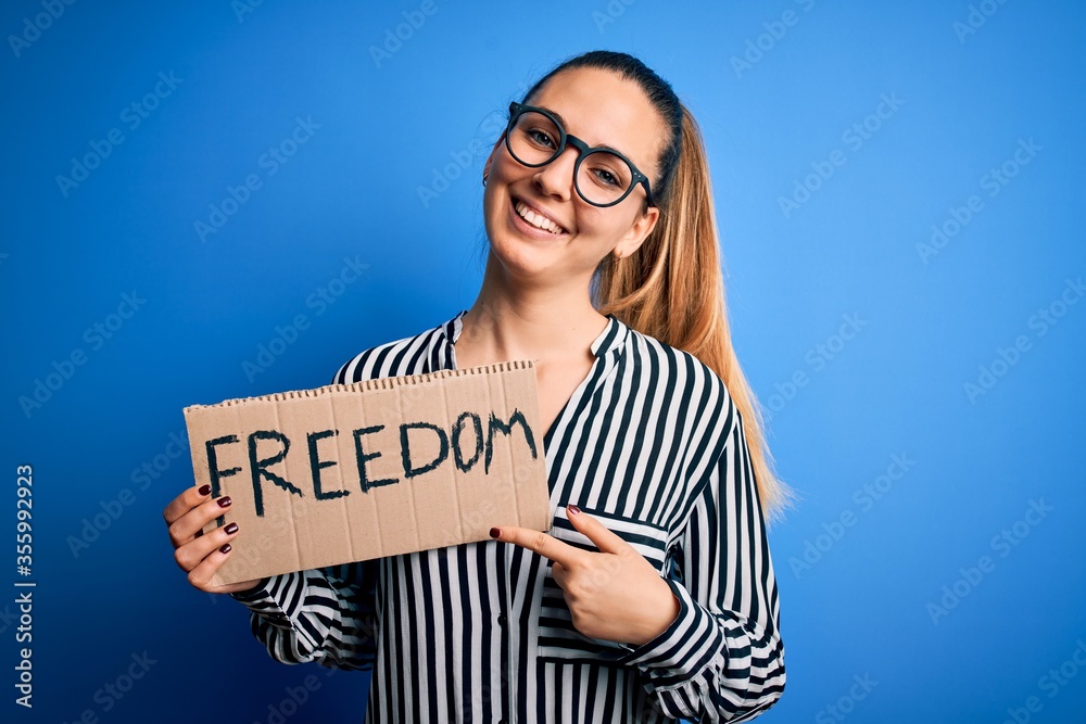 Young beautiful blonde woman with blue eyes holding banner with freedom message very happy pointing with hand and finger Young beautiful blonde woman with blue eyes holding banner with freedom message very happy pointing with hand and finger