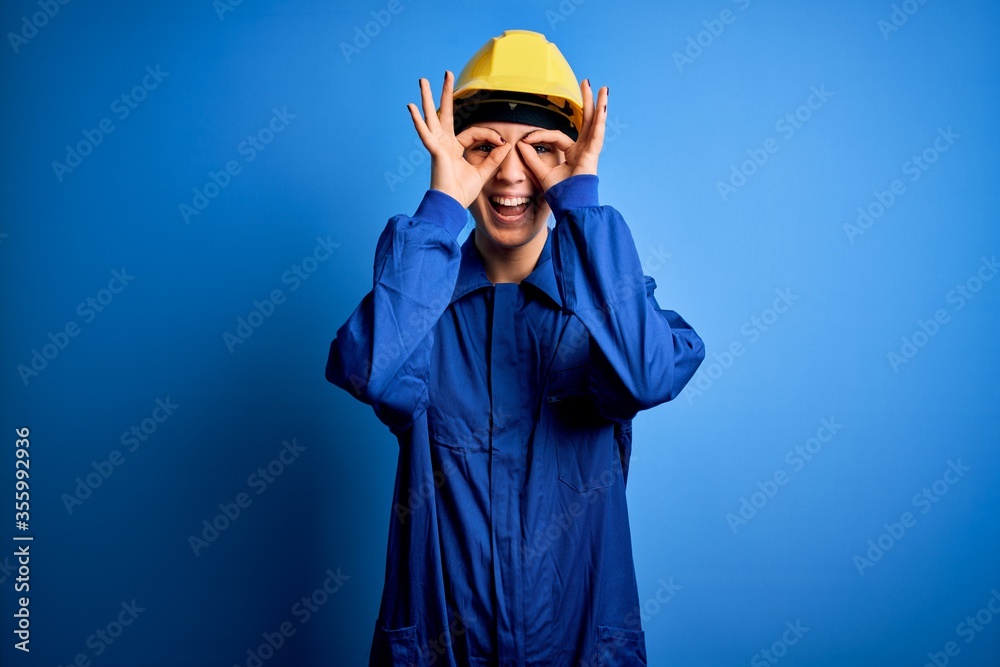 Young beautiful worker woman with blue eyes wearing security helmet and uniform doing ok gesture like binoculars sticking tongue out, eyes looking through fingers. Crazy expression. Young beautiful worker woman with blue eyes wearing security helmet and uniform doing ok gesture like binoculars sticking tongue out, eyes looking through fingers. Crazy expression.