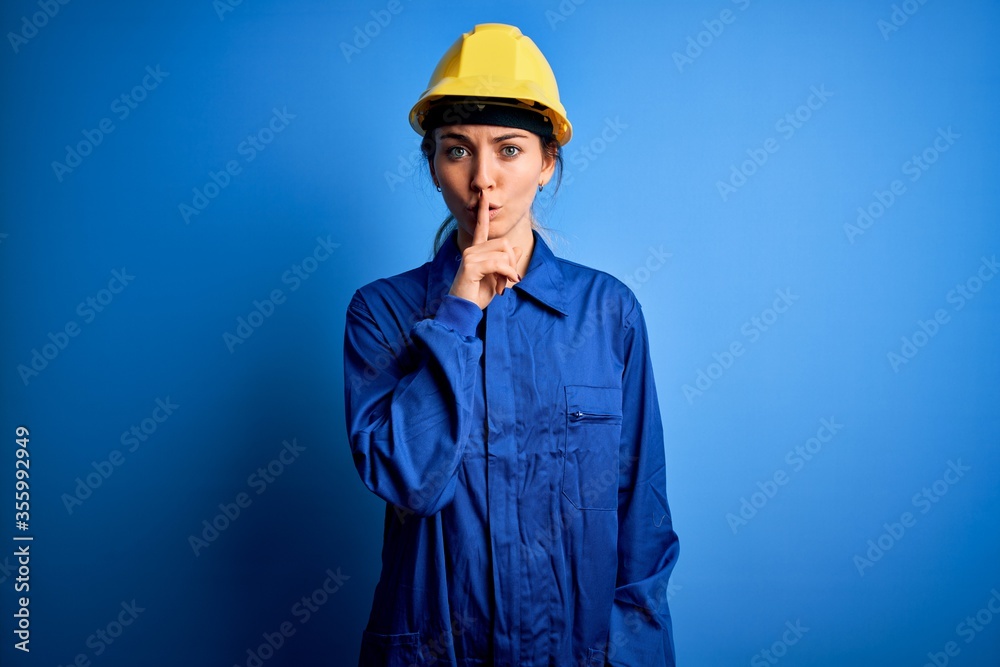 Young beautiful worker woman with blue eyes wearing security helmet and uniform asking to be quiet with finger on lips. Silence and secret concept.