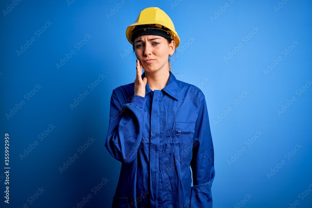 Young beautiful worker woman with blue eyes wearing security helmet and uniform touching mouth with hand with painful expression because of toothache or dental illness on teeth. Dentist