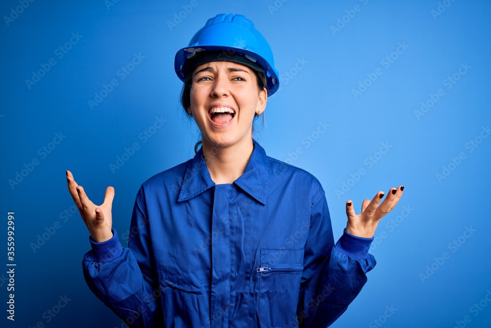 Young beautiful worker woman with blue eyes wearing security helmet and uniform crazy and mad shouting and yelling with aggressive expression and arms raised. Frustration concept. Young beautiful worker woman with blue eyes wearing security helmet and uniform crazy and mad shouting and yelling with aggressive expression and arms raised. Frustration concept.