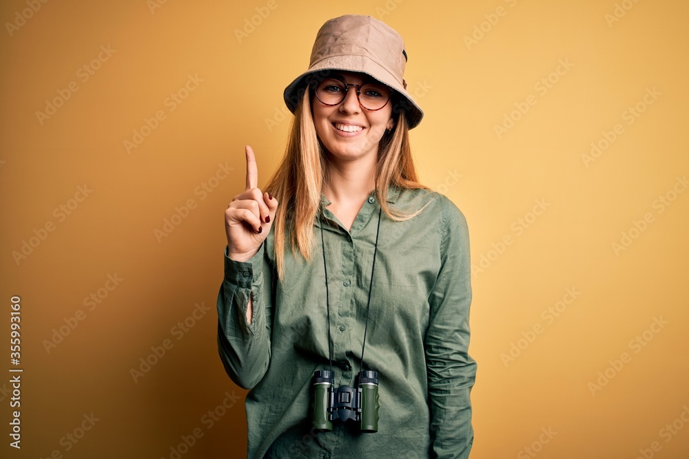 Beautiful blonde explorer woman with blue eyes wearing hat and glasses using binoculars showing and pointing up with finger number one while smiling confident and happy. Beautiful blonde explorer woman with blue eyes wearing hat and glasses using binoculars showing and pointing up with finger number one while smiling confident and happy.