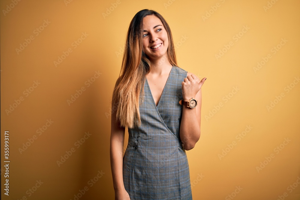 Young beautiful blonde woman with blue eyes wearing dress over yellow background smiling with happy face looking and pointing to the side with thumb up. Young beautiful blonde woman with blue eyes wearing dress over yellow background smiling with happy face looking and pointing to the side with thumb up.
