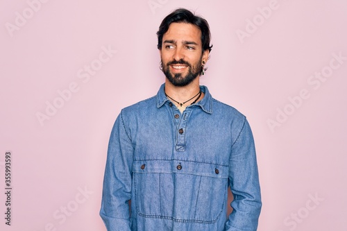 Young handsome hispanic bohemian man wearing hippie style over pink background looking away to side with smile on face, natural expression. Laughing confident.