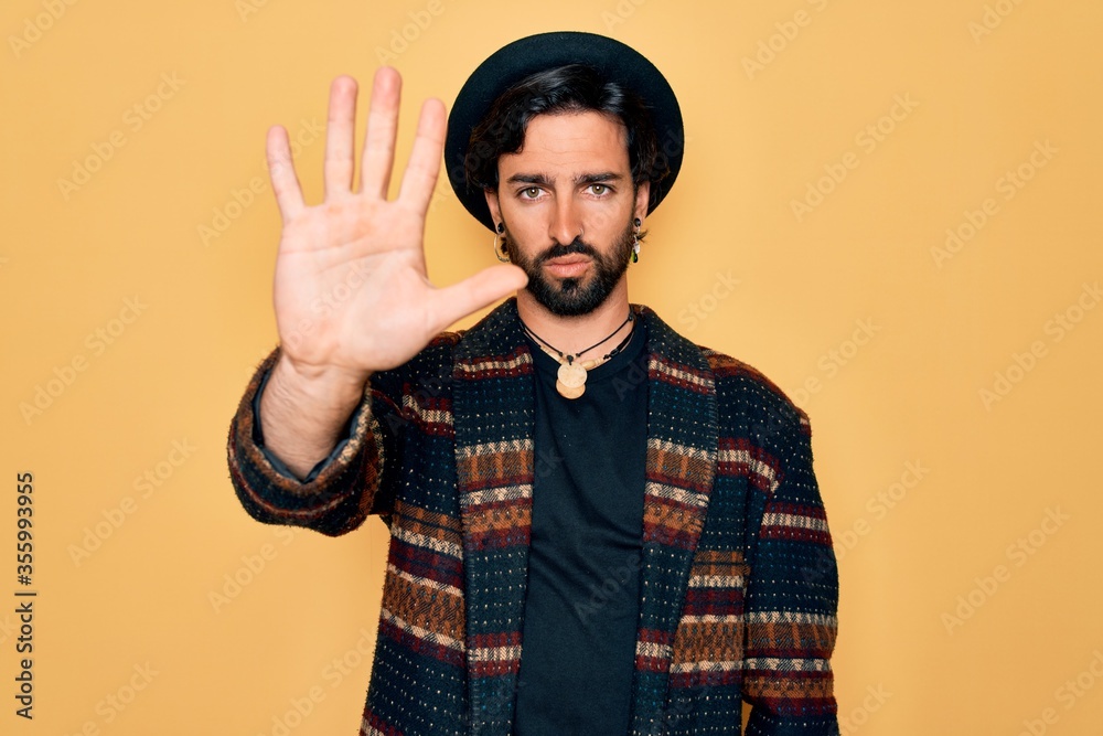 Naklejka premium Young handsome hispanic bohemian man wearing hippie style and boho hat doing stop sing with palm of the hand. Warning expression with negative and serious gesture on the face.
