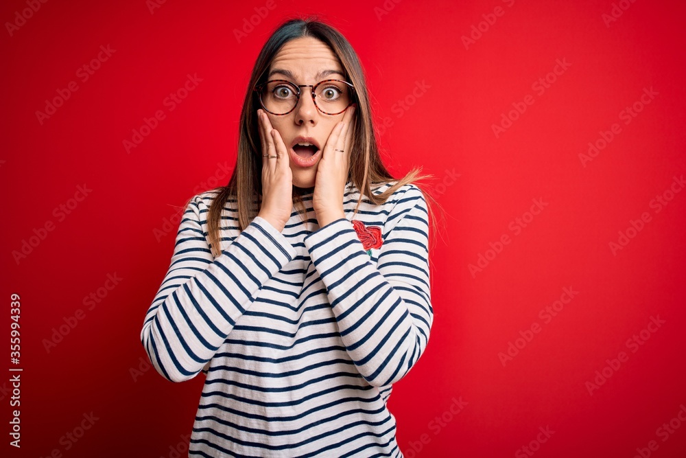 Young beautiful blonde woman with blue eyes wearing glasses standing over red background afraid and shocked, surprise and amazed expression with hands on face Young beautiful blonde woman with blue eyes wearing glasses standing over red background afraid and shocked, surprise and amazed expression with hands on face