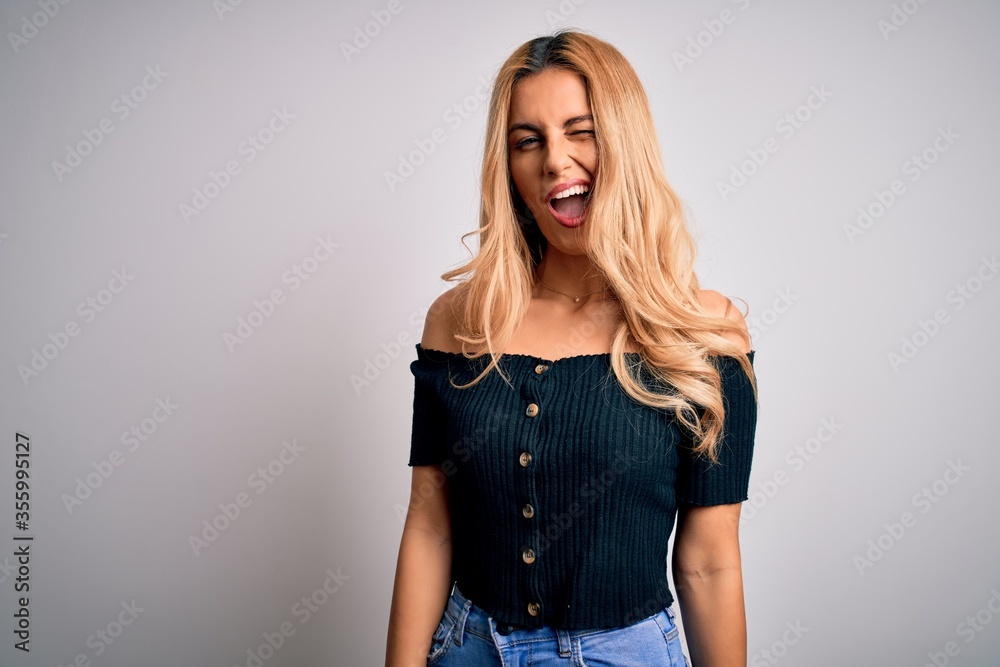 Young beautiful blonde woman wearing casual t-shirt standing over isolated white background winking looking at the camera with sexy expression, cheerful and happy face. Young beautiful blonde woman wearing casual t-shirt standing over isolated white background winking looking at the camera with sexy expression, cheerful and happy face.