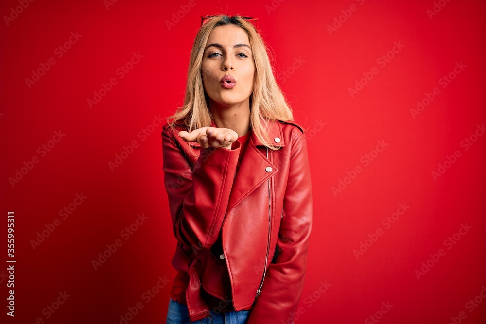 Young beautiful blonde woman wearing casual jacket standing over isolated red background looking at the camera blowing a kiss with hand on air being lovely and sexy. Love expression. Young beautiful blonde woman wearing casual jacket standing over isolated red background looking at the camera blowing a kiss with hand on air being lovely and sexy. Love expression.