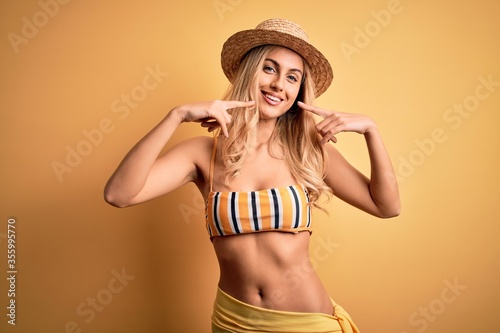 Young beautiful blonde woman on vacation wearing bikini and hat over yellow background smiling cheerful showing and pointing with fingers teeth and mouth. Dental health concept.