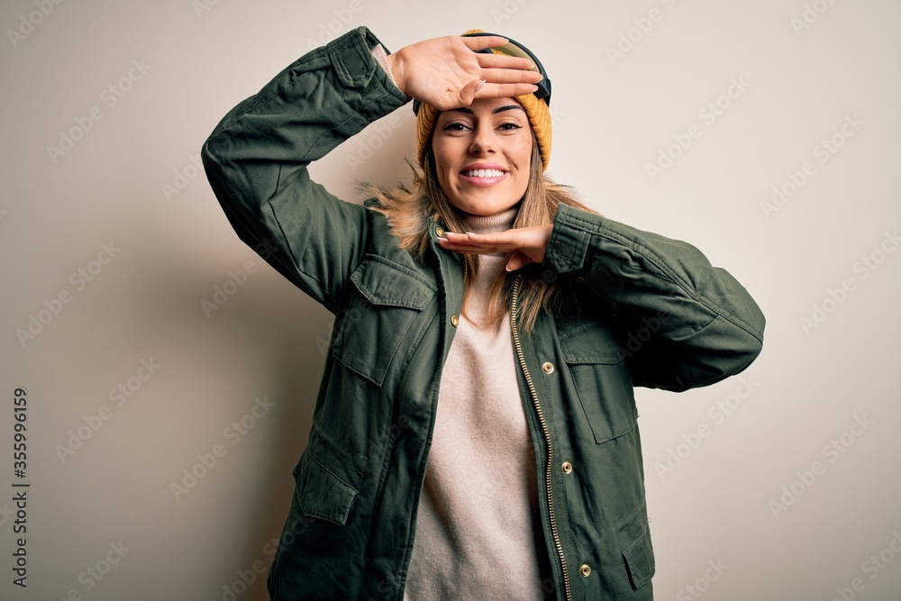 Young brunette skier woman wearing snow clothes and ski goggles over white background Smiling cheerful playing peek a boo with hands showing face. Surprised and exited