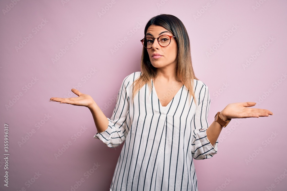 Young beautiful woman wearing casual striped t-shirt and glasses over pink background clueless and confused with open arms, no idea concept. Young beautiful woman wearing casual striped t-shirt and glasses over pink background clueless and confused with open arms, no idea concept.