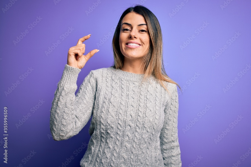 Young beautiful woman wearing casual sweater standing over isolated purple background smiling and confident gesturing with hand doing small size sign with fingers looking and the camera. Measure Young beautiful woman wearing casual sweater standing over isolated purple background smiling and confident gesturing with hand doing small size sign with fingers looking and the camera. Measure