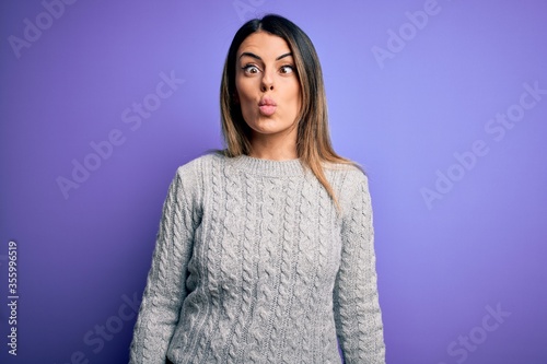 Young beautiful woman wearing casual sweater standing over isolated purple background making fish face with lips, crazy and comical gesture. Funny expression.