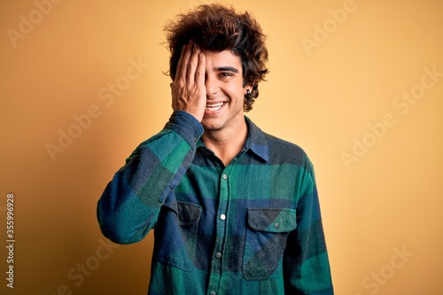Young handsome man wearing casual shirt standing over isolated yellow background covering one eye with hand, confident smile on face and surprise emotion. © Krakenimages.com