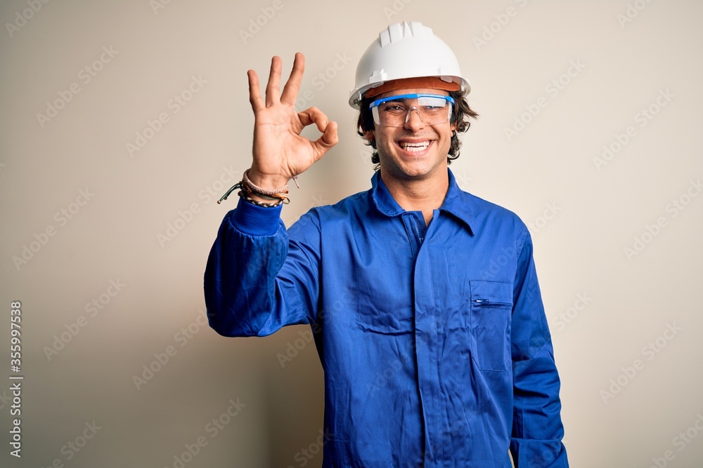 Young constructor man wearing uniform and security helmet over isolated white background smiling positive doing ok sign with hand and fingers. Successful expression.