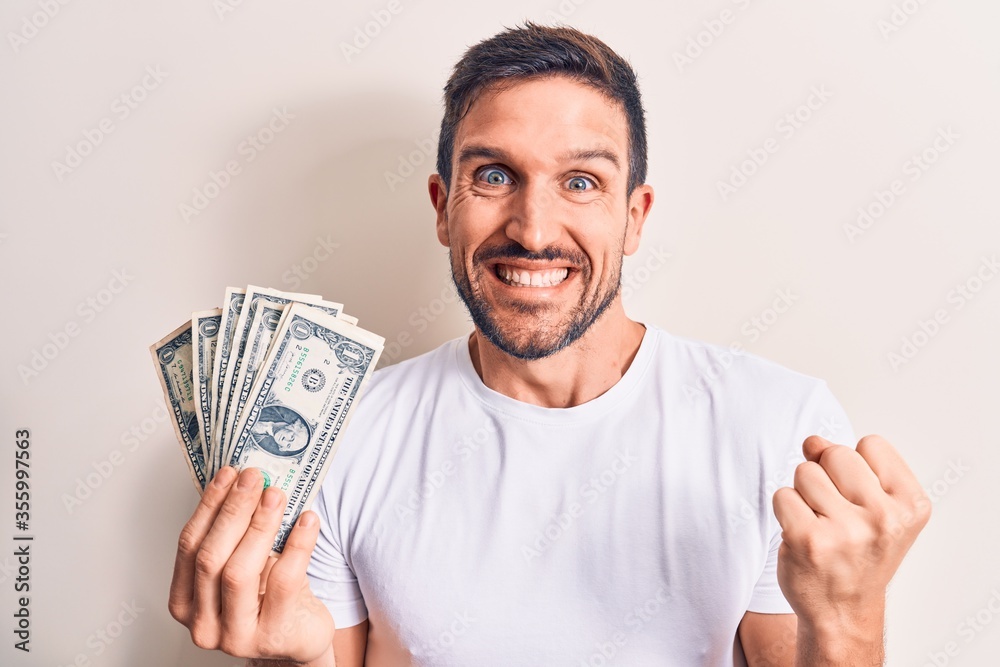 Young handsome man holding bunch of dollars banknotes over isolated white background screaming proud, celebrating victory and success very excited with raised arm Young handsome man holding bunch of dollars banknotes over isolated white background screaming proud, celebrating victory and success very excited with raised arm