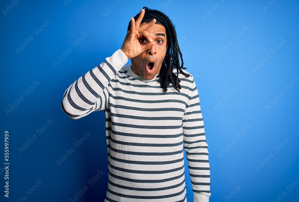 Young handsome african american afro man with dreadlocks wearing casual striped sweater doing ok gesture shocked with surprised face, eye looking through fingers. Unbelieving expression. Young handsome african american afro man with dreadlocks wearing casual striped sweater doing ok gesture shocked with surprised face, eye looking through fingers. Unbelieving expression.