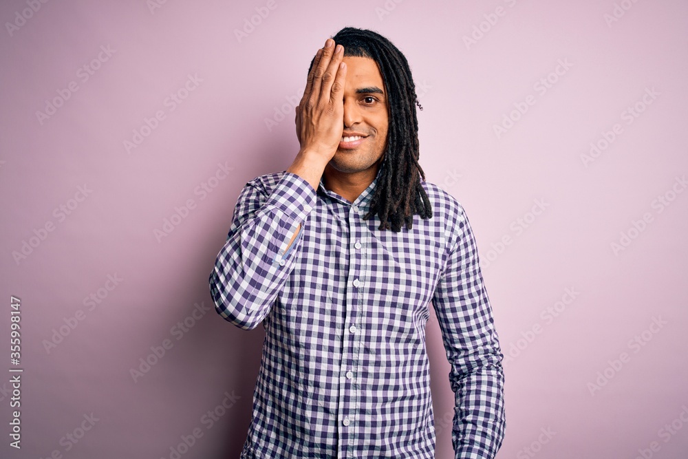 Young handsome african american afro man with dreadlocks wearing casual shirt covering one eye with hand, confident smile on face and surprise emotion. Young handsome african american afro man with dreadlocks wearing casual shirt covering one eye with hand, confident smile on face and surprise emotion.