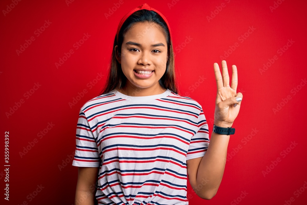 Young beautiful asian girl wearing casual striped t-shirt over isolated red background showing and pointing up with fingers number three while smiling confident and happy. Young beautiful asian girl wearing casual striped t-shirt over isolated red background showing and pointing up with fingers number three while smiling confident and happy.