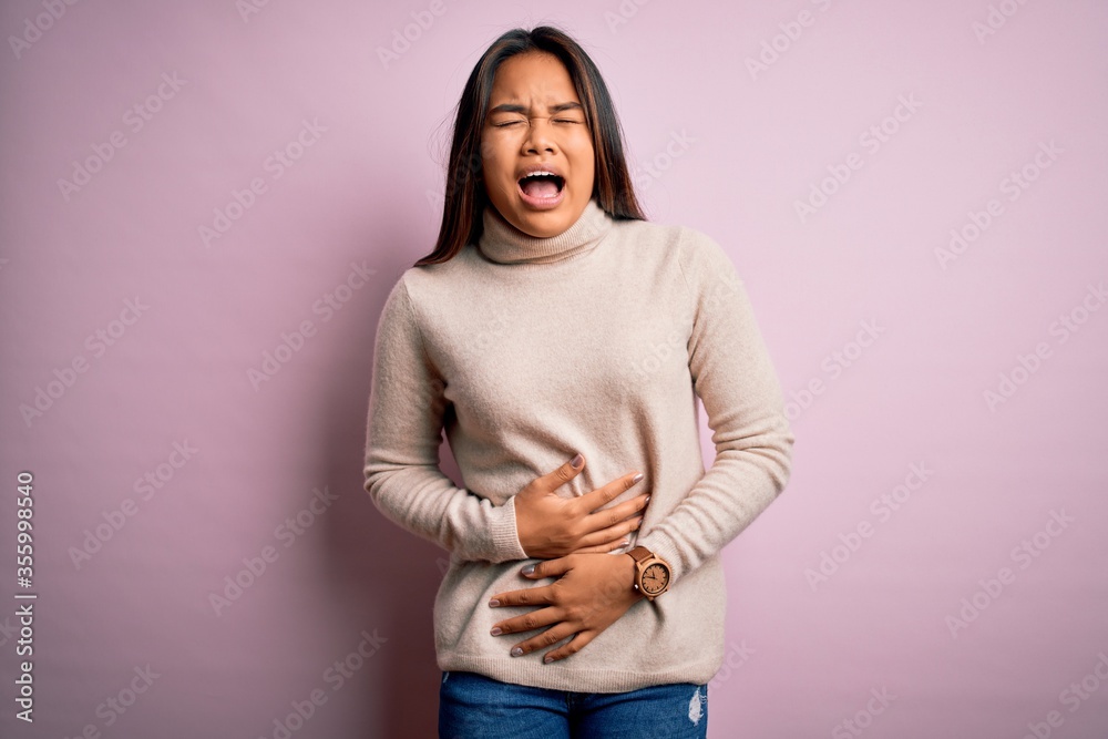 Young beautiful asian girl wearing casual turtleneck sweater over isolated pink background with hand on stomach because nausea, painful disease feeling unwell. Ache concept. Young beautiful asian girl wearing casual turtleneck sweater over isolated pink background with hand on stomach because nausea, painful disease feeling unwell. Ache concept.