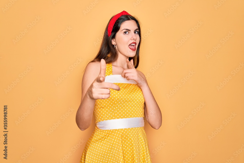 Young beautiful pin up woman wearing 50s fashion vintage dress over yellow background pointing fingers to camera with happy and funny face. Good energy and vibes. Young beautiful pin up woman wearing 50s fashion vintage dress over yellow background pointing fingers to camera with happy and funny face. Good energy and vibes.