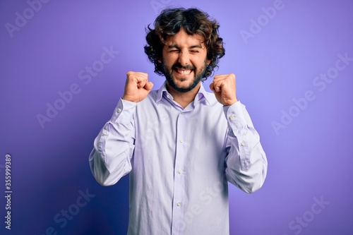 Young handsome business man with beard wearing shirt standing over purple background excited for success with arms raised and eyes closed celebrating victory smiling. Winner concept.