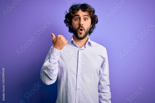 Young handsome business man with beard wearing shirt standing over purple background Surprised pointing with hand finger to the side, open mouth amazed expression.
