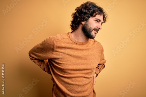 Young handsome man with beard wearing casual sweater standing over yellow background Suffering of backache, touching back with hand, muscular pain