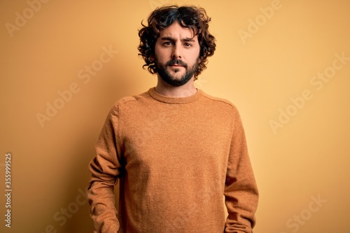 Young handsome man with beard wearing casual sweater standing over yellow background Relaxed with serious expression on face. Simple and natural looking at the camera.