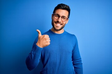 Young handsome man with beard wearing casual sweater and glasses over blue background doing happy thumbs up gesture with hand. Approving expression looking at the camera showing success.
