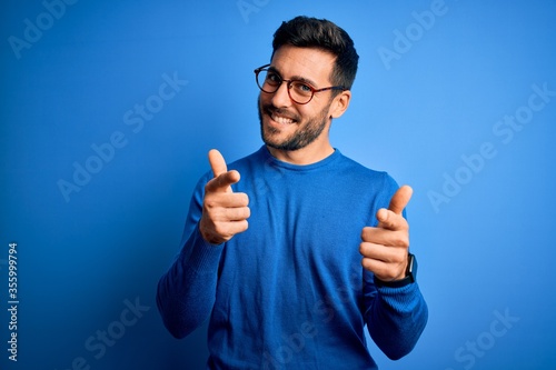 Young handsome man with beard wearing casual sweater and glasses over blue background pointing fingers to camera with happy and funny face. Good energy and vibes.