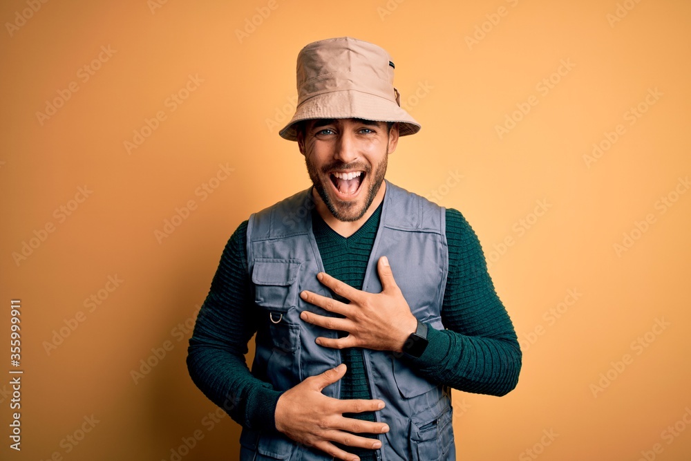 Handsome tourist man with beard on vacation wearing explorer hat over yellow background smiling and laughing hard out loud because funny crazy joke with hands on body. Handsome tourist man with beard on vacation wearing explorer hat over yellow background smiling and laughing hard out loud because funny crazy joke with hands on body.