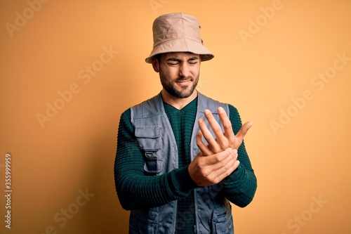 Handsome tourist man with beard on vacation wearing explorer hat over yellow background Suffering pain on hands and fingers, arthritis inflammation