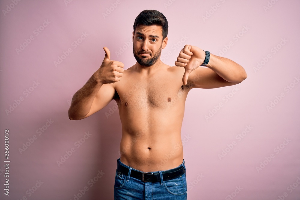 Young handsome strong man with beard shirtless standing over isolated pink background Doing thumbs up and down, disagreement and agreement expression. Crazy conflict