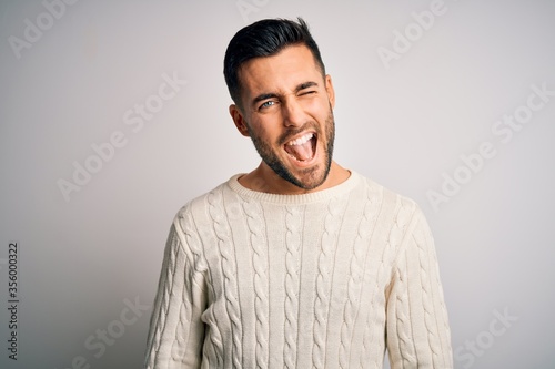 Young handsome man wearing casual sweater standing over isolated white background winking looking at the camera with sexy expression, cheerful and happy face. © Krakenimages.com