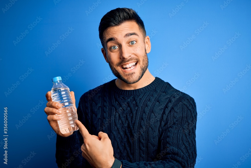 Young handsome man drinking bottle of water to refeshment over blue background very happy pointing with hand and finger Young handsome man drinking bottle of water to refeshment over blue background very happy pointing with hand and finger