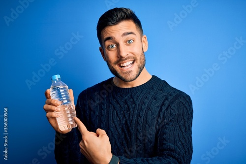 Young handsome man drinking bottle of water to refeshment over blue background very happy pointing with hand and finger