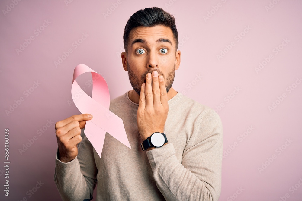 Young handsome man holding pink cancer ribbon symbol of support over isolated background cover mouth with hand shocked with shame for mistake, expression of fear, scared in silence, secret concept Young handsome man holding pink cancer ribbon symbol of support over isolated background cover mouth with hand shocked with shame for mistake, expression of fear, scared in silence, secret concept