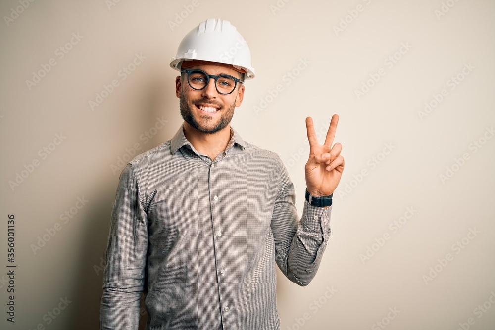 Young architect man wearing builder safety helmet over isolated background smiling with happy face winking at the camera doing victory sign. Number two. Young architect man wearing builder safety helmet over isolated background smiling with happy face winking at the camera doing victory sign. Number two.