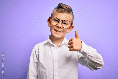 Young little caucasian kid with blue eyes wearing glasses and white shirt over purple background doing happy thumbs up gesture with hand. Approving expression looking at the camera showing success.