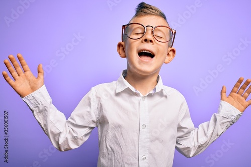 Young little caucasian kid with blue eyes wearing glasses and white shirt over purple background celebrating mad and crazy for success with arms raised and closed eyes screaming excited