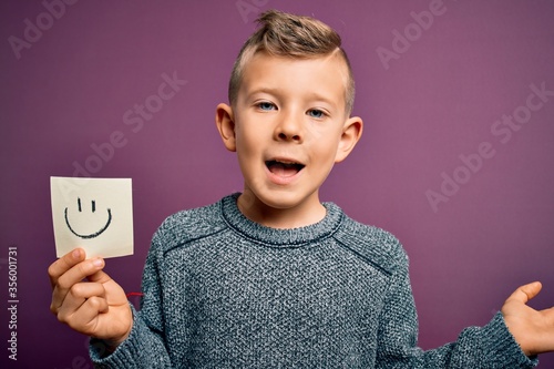 Young little caucasian kid showing smiley face on a paper note as happy message very happy and excited, winner expression celebrating victory screaming with big smile and raised hands
