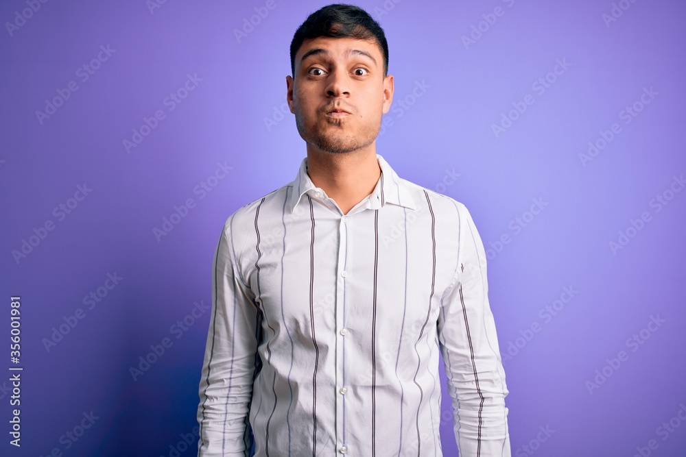 Obraz premium Young handsome hispanic man wearing elegant business shirt standing over purple background puffing cheeks with funny face. Mouth inflated with air, crazy expression.